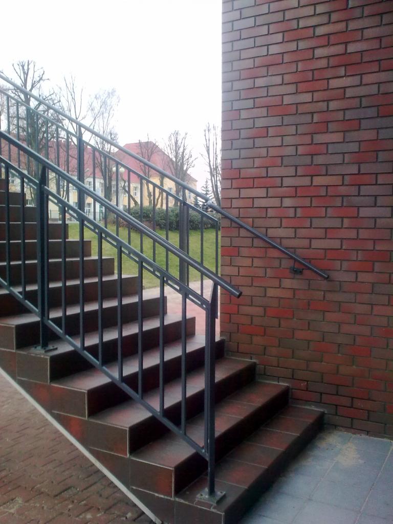 Zewnętrzne schody wyłożone brązowymi płytkami, z czarną metalową balustradą, przylegające do ściany z czerwonej cegły.