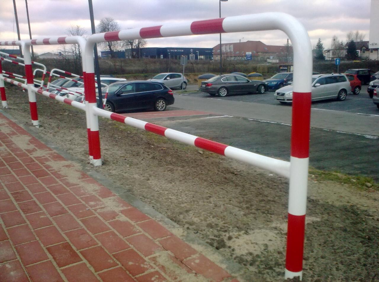 Barierki chodnikowe z rur stalowych, pomalowane w biało-czerwone pasy, oddzielające chodnik z czerwonej kostki brukowej od parkingu z samochodami w tle.