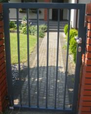 Ciemnoszara furtka z metalowych prętów, zamontowana między ceglanymi słupkami, prowadząca do ścieżki w ogrodzie.