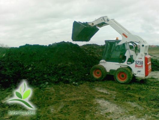 Biała miniładowarka Bobcat S250 z podniesioną łyżką nad stertą ciemnej ziemi na błotnistym terenie, logo InterGarden w lewym dolnym rogu.
