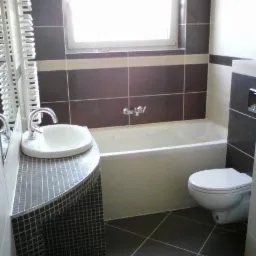 Wykończona łazienka z brązowymi płytkami, białą wanną i umywalką na szafce z mozaiki. Ukończony remont łazienki z widocznymi elementami wyposażenia.