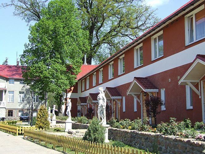 Budynek wielorodzinny z czerwonym dachem i rzędem białych statuet w ogrodzie, widoczny fragment drugiego budynku z czerwonym dachem i zaparkowanym samochodem.