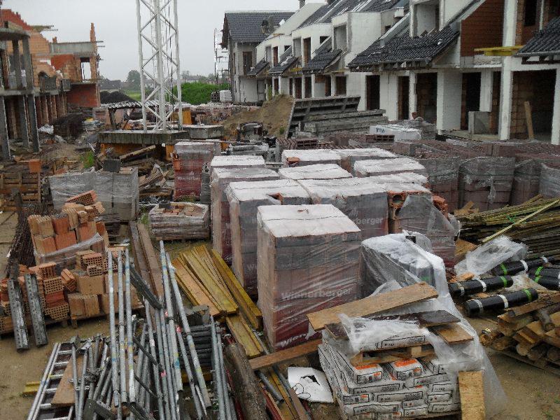 Plac budowy z widocznymi budynkami w stanie surowym, paletami cegieł marki Wienerberger, deskami szalunkowymi i rusztowaniami, na tle pochmurnego nieba.