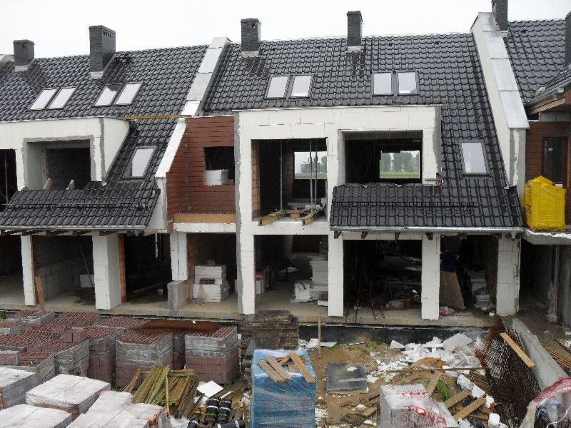 Wznoszenie szeregu budynków jednorodzinnych w stanie surowym, widoczne czarne dachówki, kominy i okna dachowe, ściany z białego pustaka, cegły i niezabezpieczone obróbki blacharskie, teren budowy...