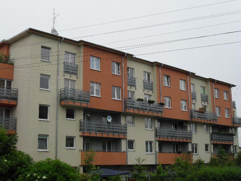 Nowoczesny budynek wielorodzinny z balkonami, pomalowany na beżowo i pomarańczowo, widoczna antena satelitarna na jednym z balkonów, w tle pochmurne niebo i zieleń drzew.