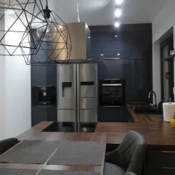 Nowoczesna kuchnia z ciemnymi, lakierowanymi szafkami, drewnianym blatem i stalowymi sprzętami AGD, oświetlona geometrycznymi lampami wiszącymi, uzupełniona owocami na blacie.