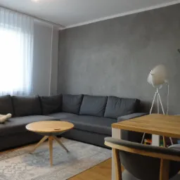 Szary narożnik z pluszowym misiem pod oknem, okrągły stolik kawowy na wzorzystym dywanie, drewniany stół z krzesłami w tle, ściana w odcieniach szarości, lampa na trójnogu i komoda z żółtą ścianką...