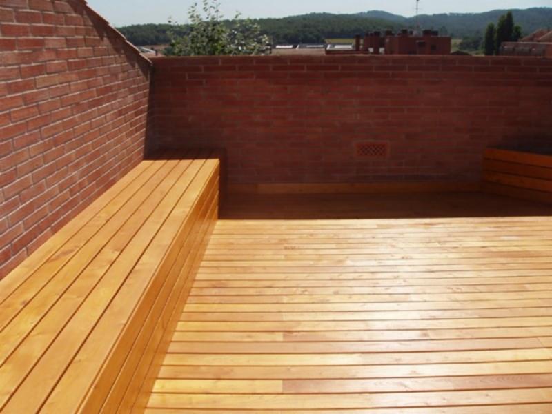 Drewniany taras na dachu z ławką z tego samego drewna, obok ceglany mur.