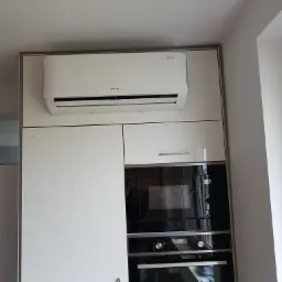 Biały klimatyzator marki Rotenso zamontowany nad zabudową kuchenną z piekarnikiem i mikrofalówką, widok na ścianę i sufit.