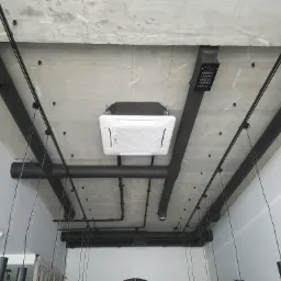 Kasetonowa jednostka klimatyzacyjna zamontowana na surowym, betonowym suficie z widocznymi czarnymi rurami wentylacyjnymi i elementami oświetlenia w nowoczesnym biurze.