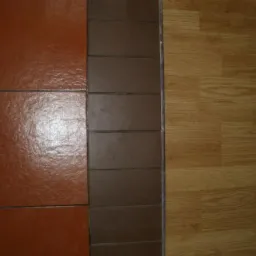 Połączenie brązowych płytek podłogowych, ciemnobrązowej listwy i paneli podłogowych imitujących drewno, widoczne z góry.