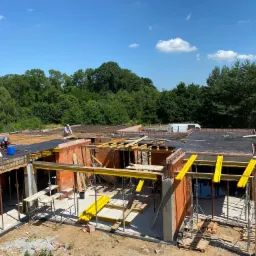 Wznoszenie ścian parterowego budynku z czerwonej cegły, widoczne ż&oacute;łte podpory stropu, pracownicy w trakcie montażu zbrojenia stropu, w tle zieleń drzew i błękitne niebo.