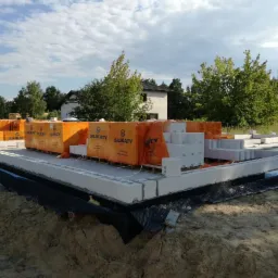 Fundamenty domu w trakcie budowy, zabezpieczone czarną folią, obok palety z bloczkami silikatowymi w pomarańczowych opakowaniach, w tle budynek w stanie surowym otoczony zielenią.