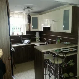 Nowoczesna kuchnia z barkiem, wykończona w odcieniach brązu i bieli, z chromowanymi stołkami barowymi i płytkami podłogowymi w odcieniach beżu. Widoczne okno z firanką i dekoracyjna lampa sufitowa.