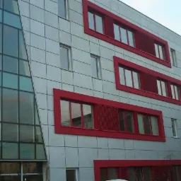 Nowoczesny budynek biurowy z szarą elewacją w płytach i czerwonymi obramowaniami okien oraz przeszkloną fasadą po lewej stronie.