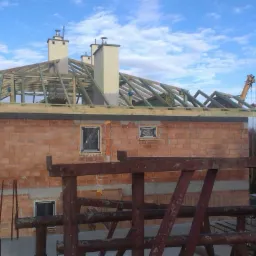 Budowa domu z cegły: ściany wzniesione, dach w trakcie konstrukcji z widocznym drewnianym szkieletem i kominami. Żuraw budowlany w tle.