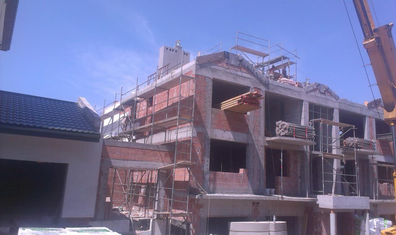 Budowa domu murowanego w trakcie realizacji: widoczne ściany z cegły, rusztowania i elementy konstrukcyjne. Na pierwszym planie fragment dachu i materiały budowlane.