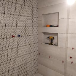 Danak - Układanie płytek w łazience: ściana z geometrycznym wzorem, druga ściana z wnękami i poziomą strukturą, widoczne narzędzia glazurnicze i krzyżyki dystansowe.