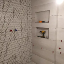 Układanie płytek w łazience: ściana z geometrycznym wzorem, druga ściana z wnękami i poziomą strukturą, widoczne narzędzia glazurnicze i krzyżyki dystansowe.