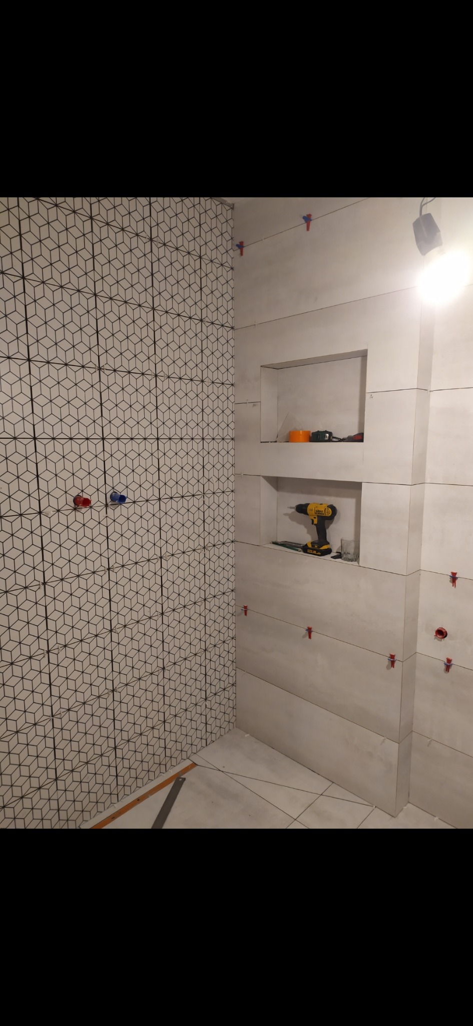 Układanie płytek w łazience: ściana z geometrycznym wzorem, druga ściana z wnękami i poziomą strukturą, widoczne narzędzia glazurnicze i krzyżyki dystansowe.