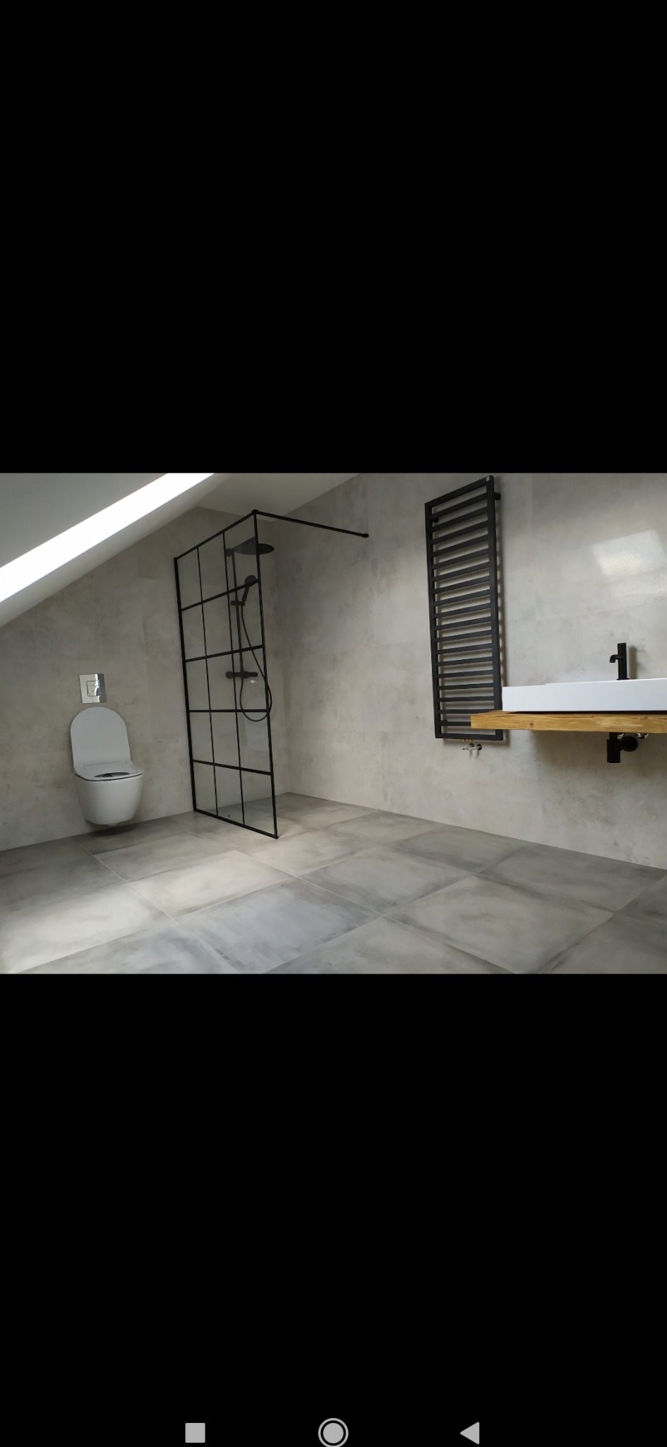 Nowoczesna łazienka na poddaszu z szarymi płytkami, kabiną prysznicową z czarnymi profilami, wiszącą toaletą i grzejnikiem drabinkowym w kolorze czarnym.