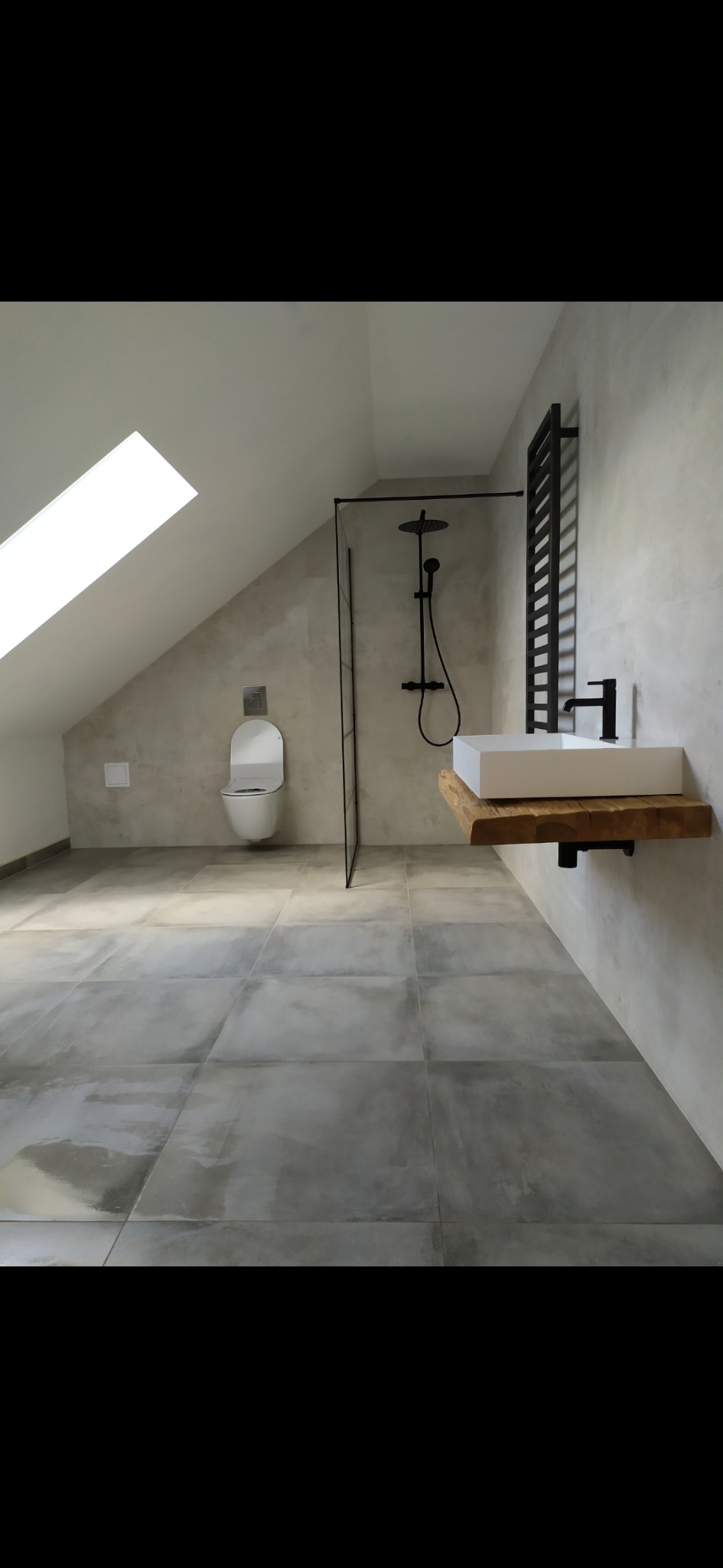 Nowoczesna łazienka na poddaszu z szarą, wielkoformatową terakotą, kabiną prysznicową typu walk-in z czarną armaturą i umywalką nablatową na blacie z surowego drewna.