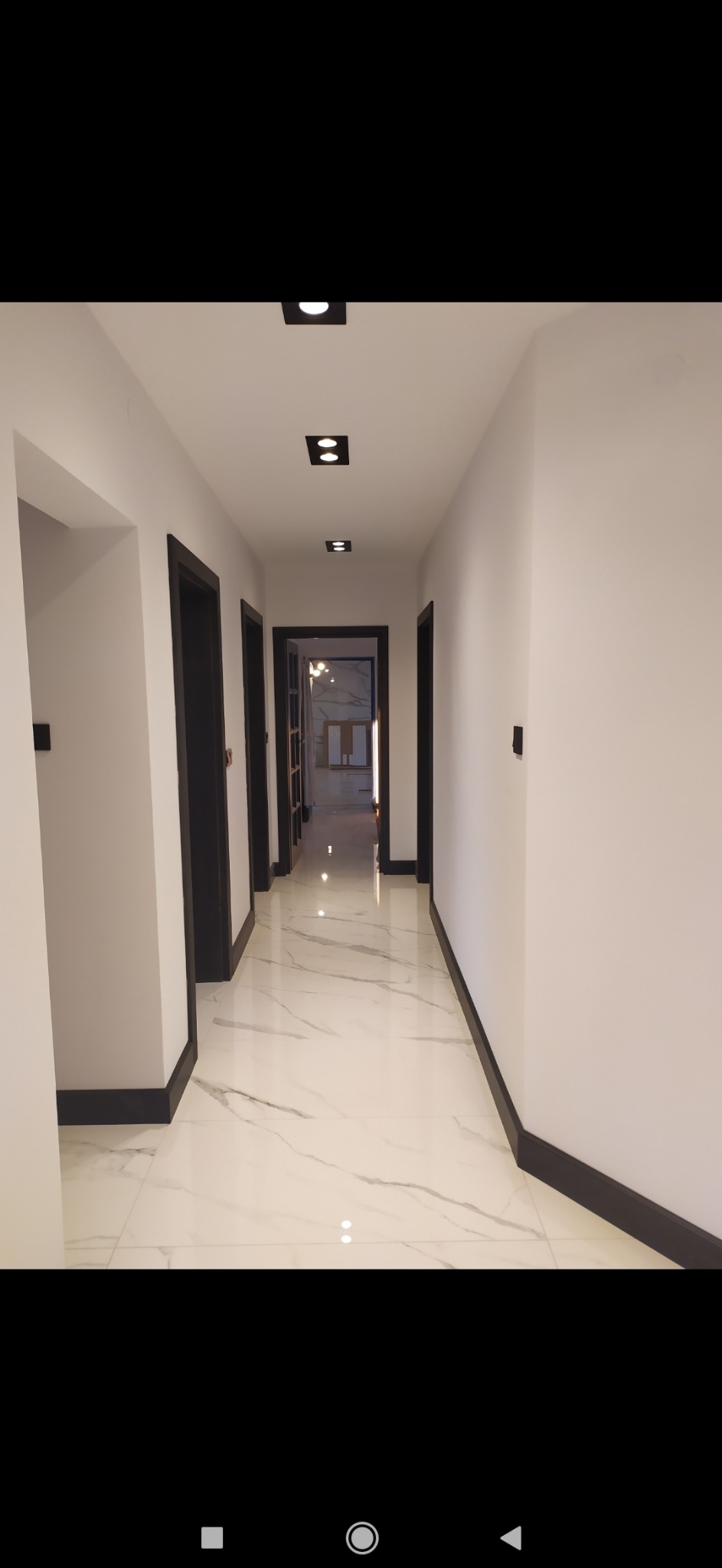 Długi korytarz z białymi ścianami, marmurową podłogą i czarnymi listwami przypodłogowymi oraz obramowaniami drzwi, oświetlony czarnymi, kwadratowymi lampami sufitowymi.