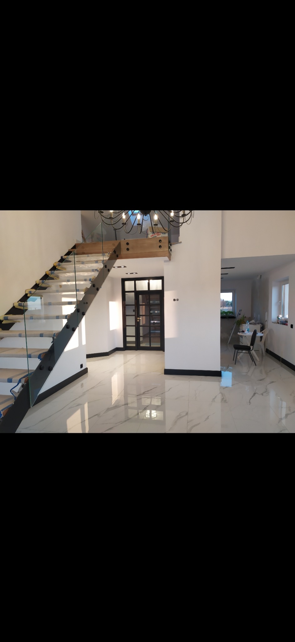 Wnętrze domu w Przeworsku z marmurową podłogą, szklanymi schodami z drewnianymi stopniami zabezpieczonymi folią i czarną stolarką drzwiową, w trakcie wykańczania.