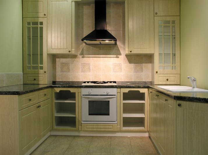 Jasna, klasyczna zabudowa kuchenna z drewnianymi frontami, czarnym blatem i czarnym okapem, płytki ceramiczne nad blatem, widoczna płyta grzewcza i piekarnik.