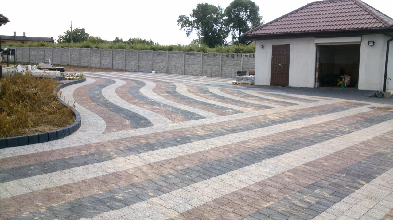 Ułożona kostka brukowa tworząca wzór falujących pasów w odcieniach szarości i brązu na podjeździe. W tle widoczny budynek gospodarczy i betonowe ogrodzenie.