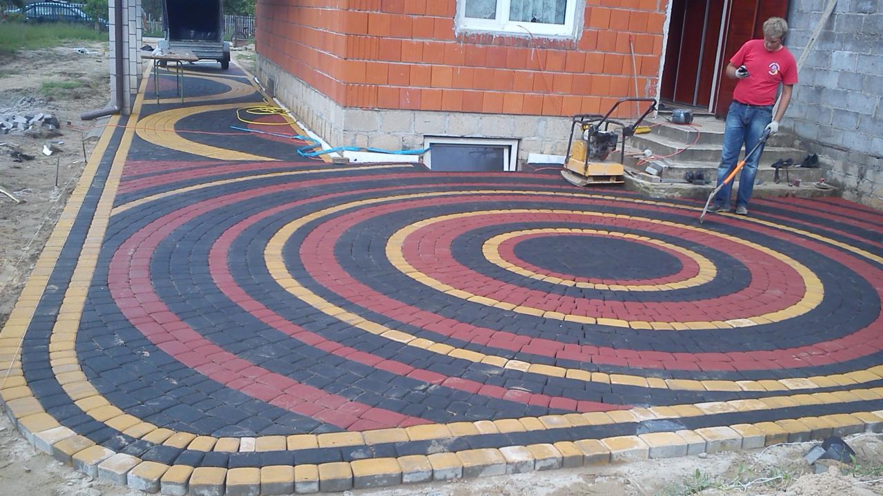Okrągły wzór z kostki brukowej w odcieniach czerni, czerwieni i żółci, obok budynku z cegły i pracownik z narzędziem. Widoczna ubijarka do kostki.