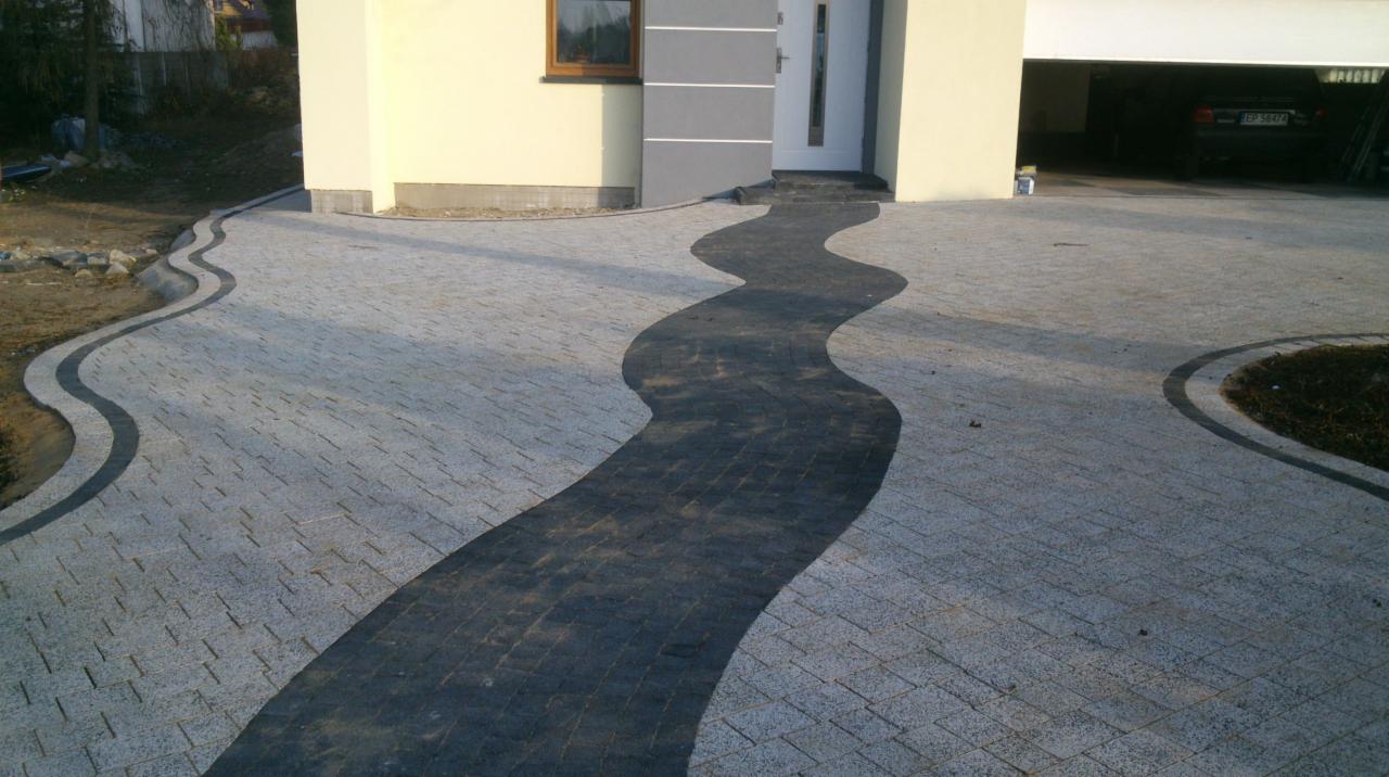 Falista ścieżka z kostki brukowej w odcieniach szarości prowadząca do domu z garażem. Dekoracyjne obramowanie z ciemnej kostki. Widok z góry.