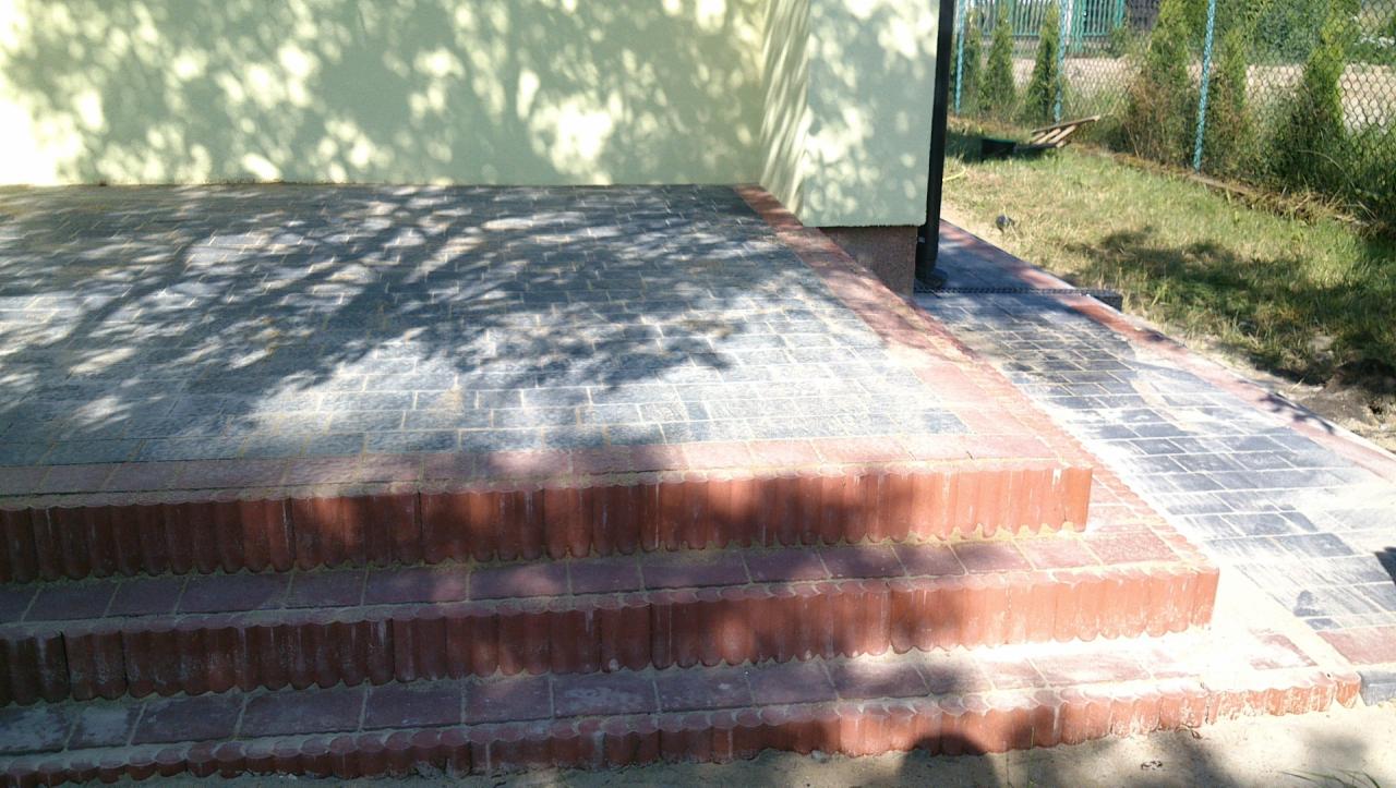 Ułożona kostka brukowa na schodach wejściowych, z cieniowanymi wzorami na powierzchni i obrzeżami z cegły, prowadząca do budynku z zielonym tłem.