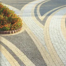 Wzór z kostki brukowej w odcieniach szarości i beżu, układający się w łukowate pasy; rabata z pomarańczowymi kwiatami w tle.