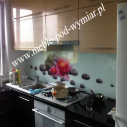 Jasne, lakierowane szafki kuchenne z poziomymi uchwytami, czarny blat, panel szklany z motywem lotosu i kamieni, kuchenka z garnkami i patelniami.