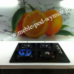 Czarny gazowy panel kuchenny z jednym palnikiem włączonym, zamontowany na białym blacie, z tłem przedstawiającym grafikę pomarańczy na marmurowej powierzchni.