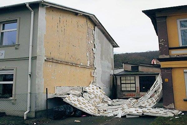 Uszkodzona elewacja budynku z odpadającym styropianem i odsłoniętym murem, widoczne ślady zawilgocenia i odpadająca farba.