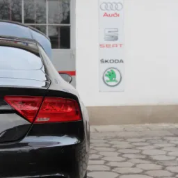 Czarny samochód Audi A7 z widocznymi tylnymi światłami i rurą wydechową, zaparkowany przed budynkiem z logo marek Volkswagen Group: VW, Audi, Seat i Skoda.