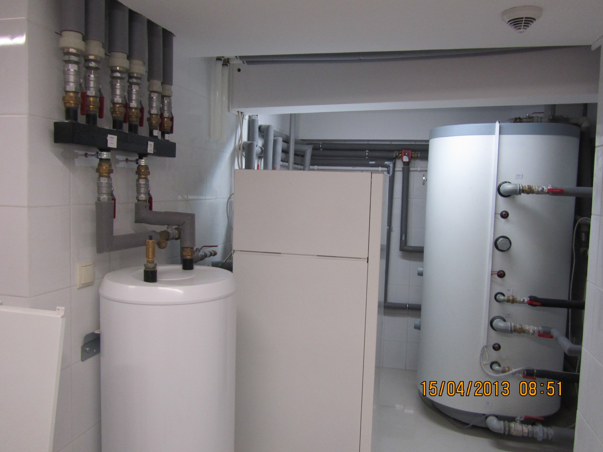 Białe zbiorniki i rury w pomieszczeniu kotłowni z płytkami, widoczne podłączenia i zawory, instalacja grzewcza.