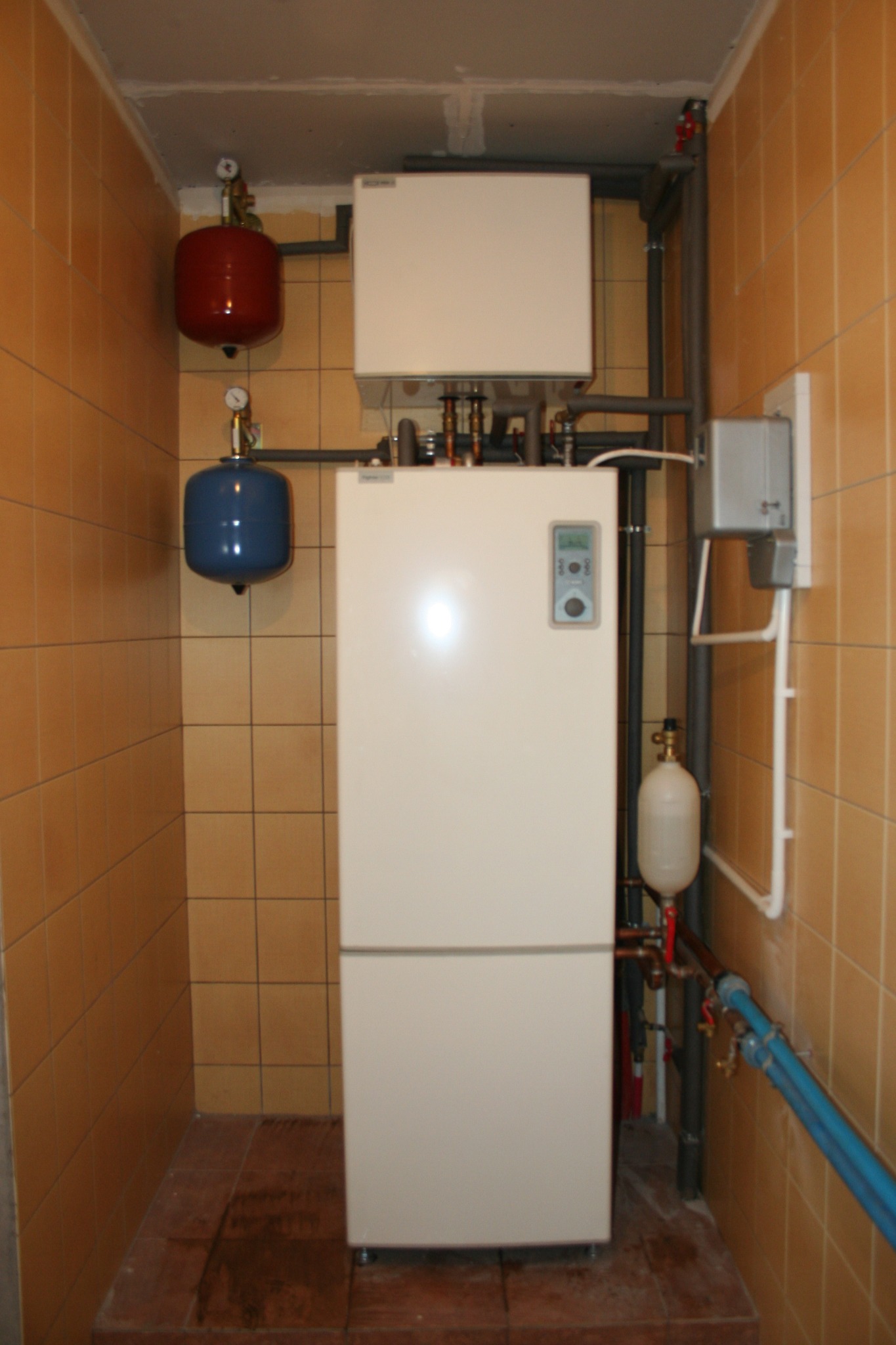 Biały, dwuczęściowy system grzewczy z czerwonym i niebieskim zbiornikiem wyrównawczym, zamontowany w pomieszczeniu z brązowymi płytkami.