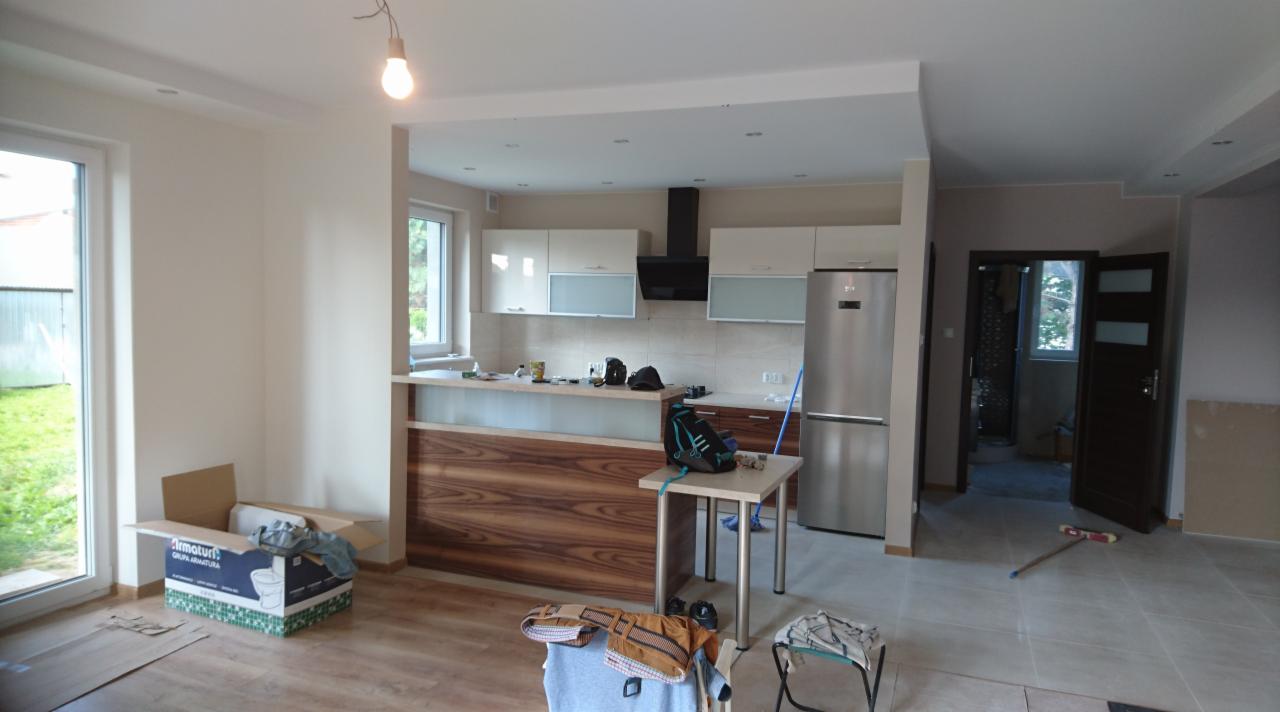 Wnętrze w trakcie prac wykończeniowych: widoczna kuchnia z zabudową, płytki na ścianach, podłoga częściowo wyłożona panelami, karton z armaturą sanitarną, oraz otwarte drzwi do innego pomieszczenia.