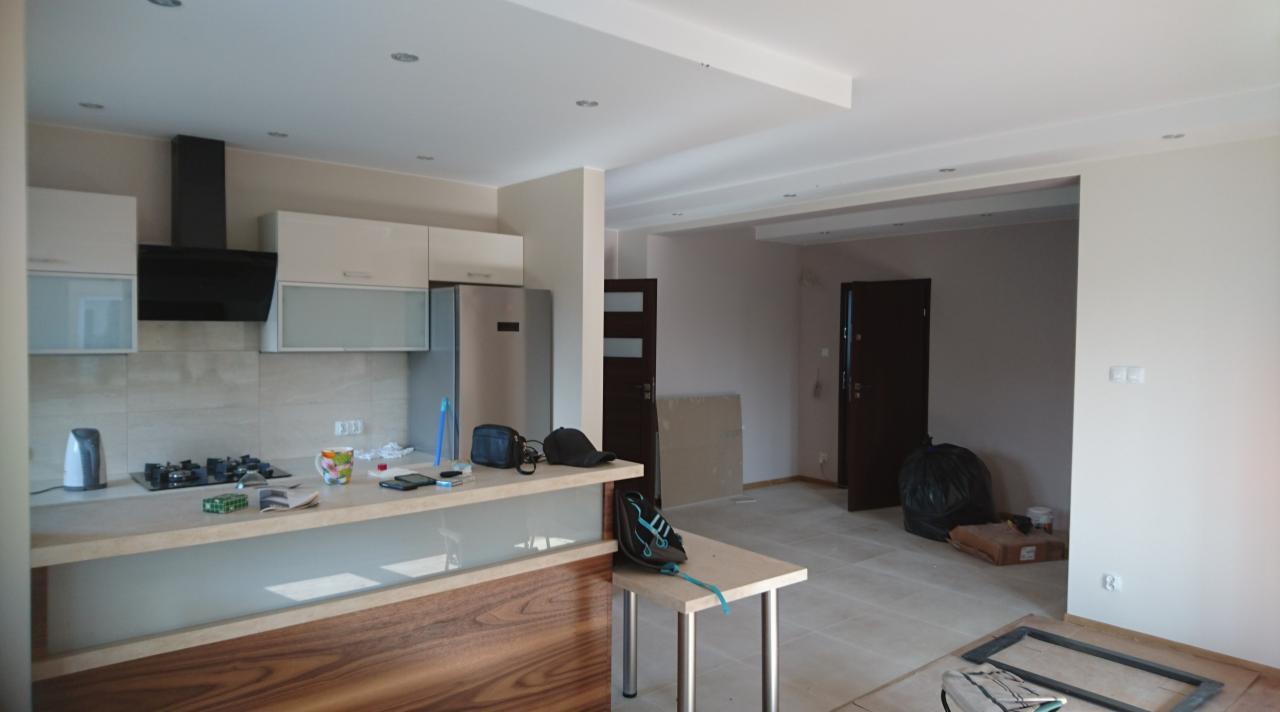 Wnętrze w trakcie prac wykończeniowych: widok na kuchnię z wyspą, fragment salonu z dwiema ciemnymi drzwiami, widoczne narzędzia i materiały budowlane, surowy stan podłogi.