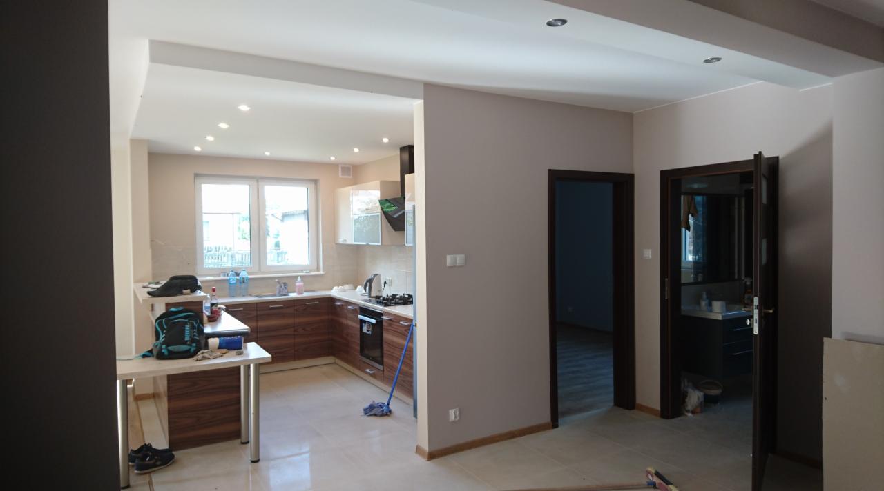 Otwarta przestrzeń: kuchnia z drewnianymi frontami szafek, widok na łazienkę przez otwarte drzwi, na blacie plecak i buty, mop oparty o szafkę.