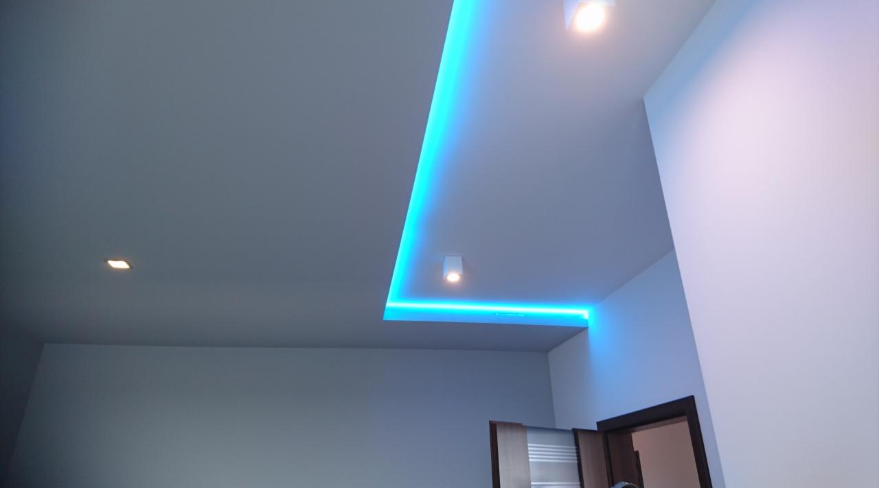 Nowoczesny sufit podwieszany z oświetleniem LED w kolorze turkusowym, widoczne halogenowe punkty świetlne i fragment otwartych drzwi.