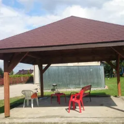 Drewniana altana z brązowym dachem z gontu bitumicznego, ustawiona na betonowej podstawie. W tle widoczny samochód dostawczy, basen i zieleń ogrodu.