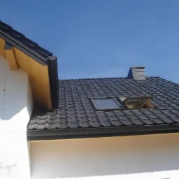 Fragment domu z czarnym dachem, otwartym oknem dachowym i kominem na tle błękitnego nieba. Widoczna biała ściana budynku z elementami drewnianymi.