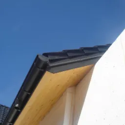 Czarne rynny dachowe zamontowane na drewnianej konstrukcji dachu, widoczne na tle błękitnego nieba i fragmentu białej ściany budynku.
