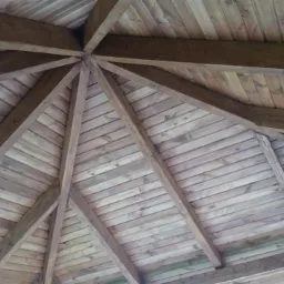 Drewniany dach pergoli, widok od dołu. Konstrukcja z belek i desek, naturalne oświetlenie, widoczne słoje drewna.