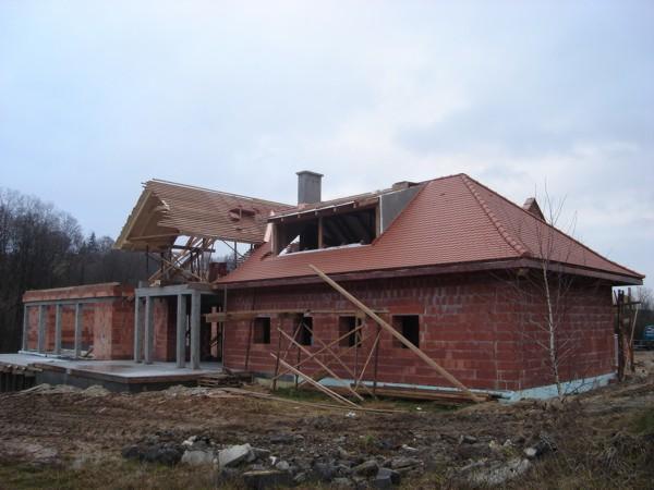 Budowa domu z czerwonej cegły: widok dachu z czerwoną dachówką i fragmentem więźby dachowej w surowym stanie. Tło: drzewa i pochmurne niebo.