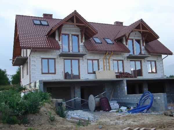 Dwukondygnacyjny dom w trakcie budowy z czerwonym dachem i drewnianymi elementami konstrukcyjnymi. Widoczny fundament i materiały budowlane.